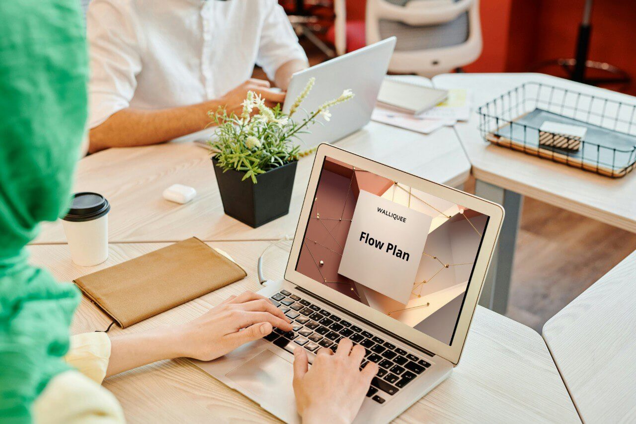 Person using a laptop with a 'Flow Plan' document on a desk with another person and office supplies.